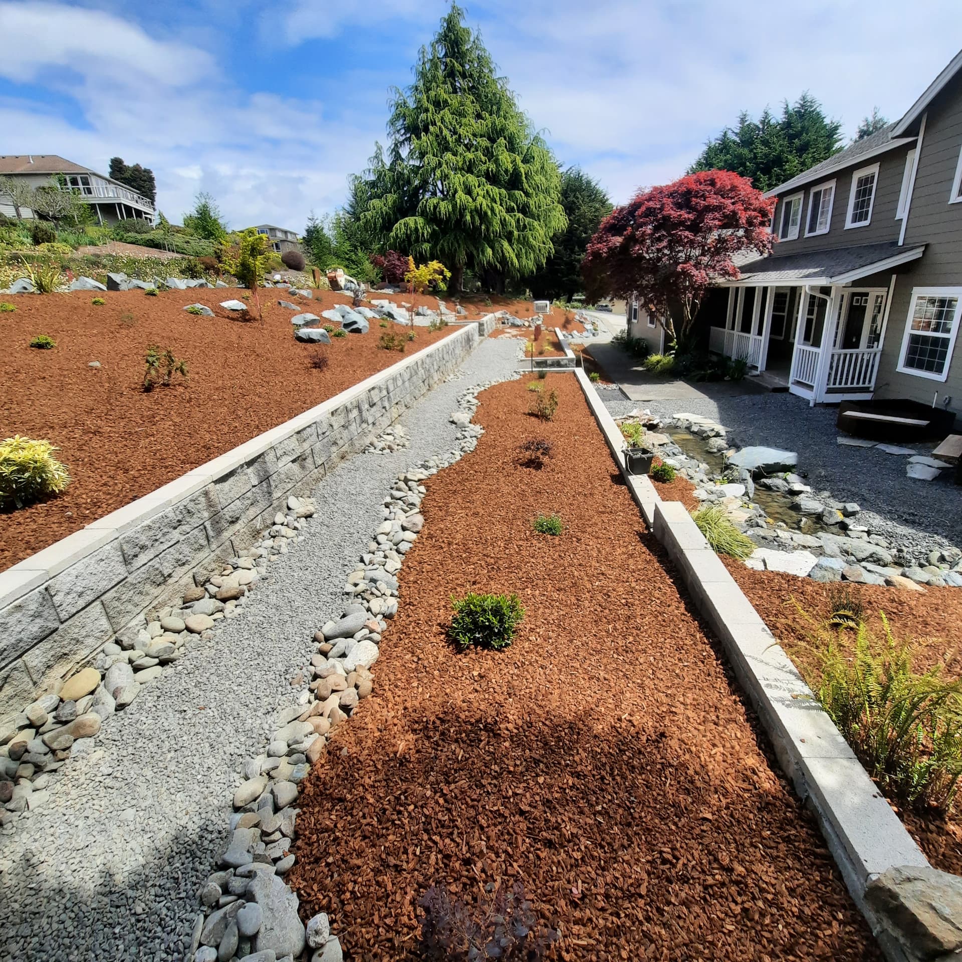 Broader view of the finished renovation with capped retaining walls, dry creek gravel, mulched beds, a pondless water feature, and reworked walkways to the house