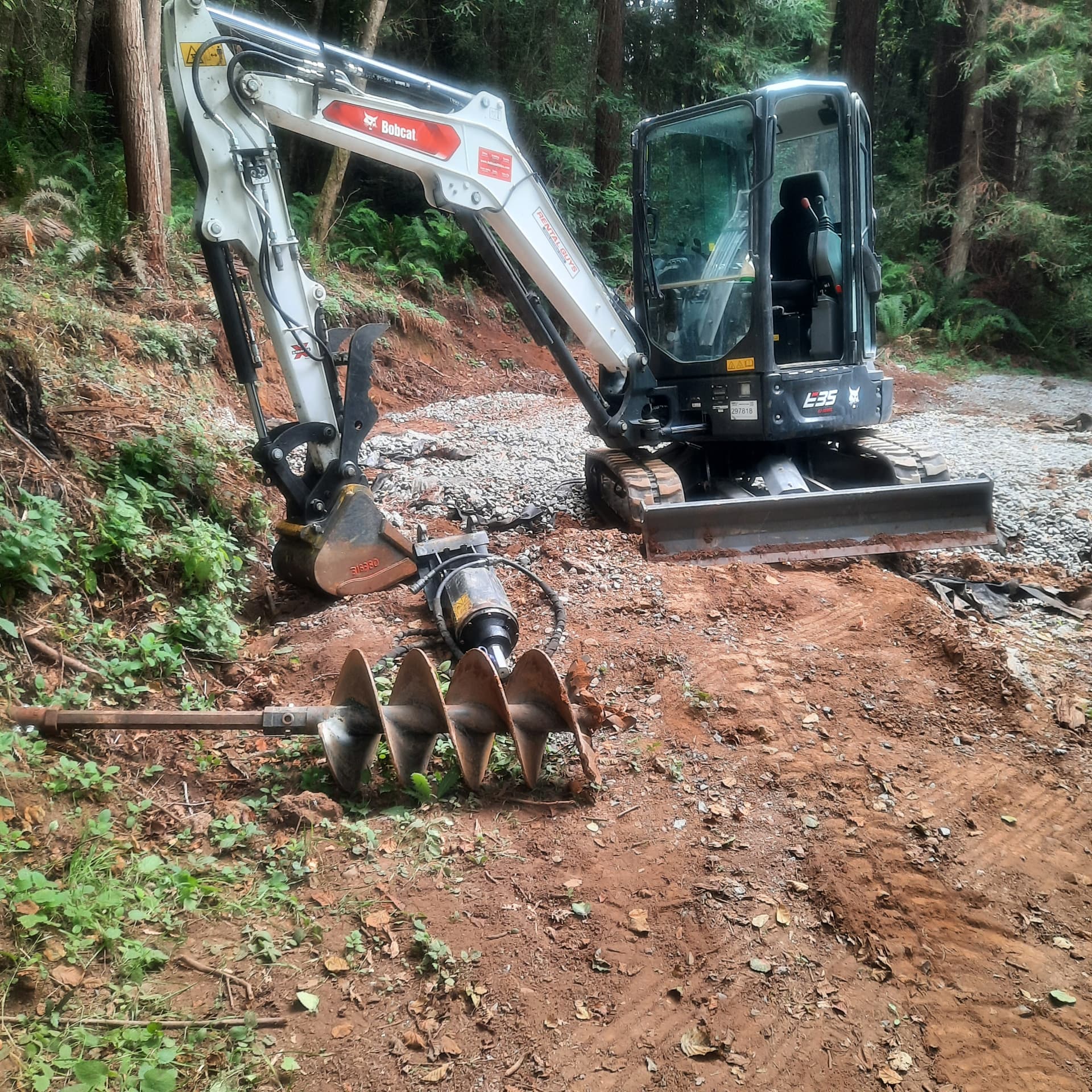 A compact excavator with an auger drive and a set of auger bits staged on a forested Humboldt County job site.