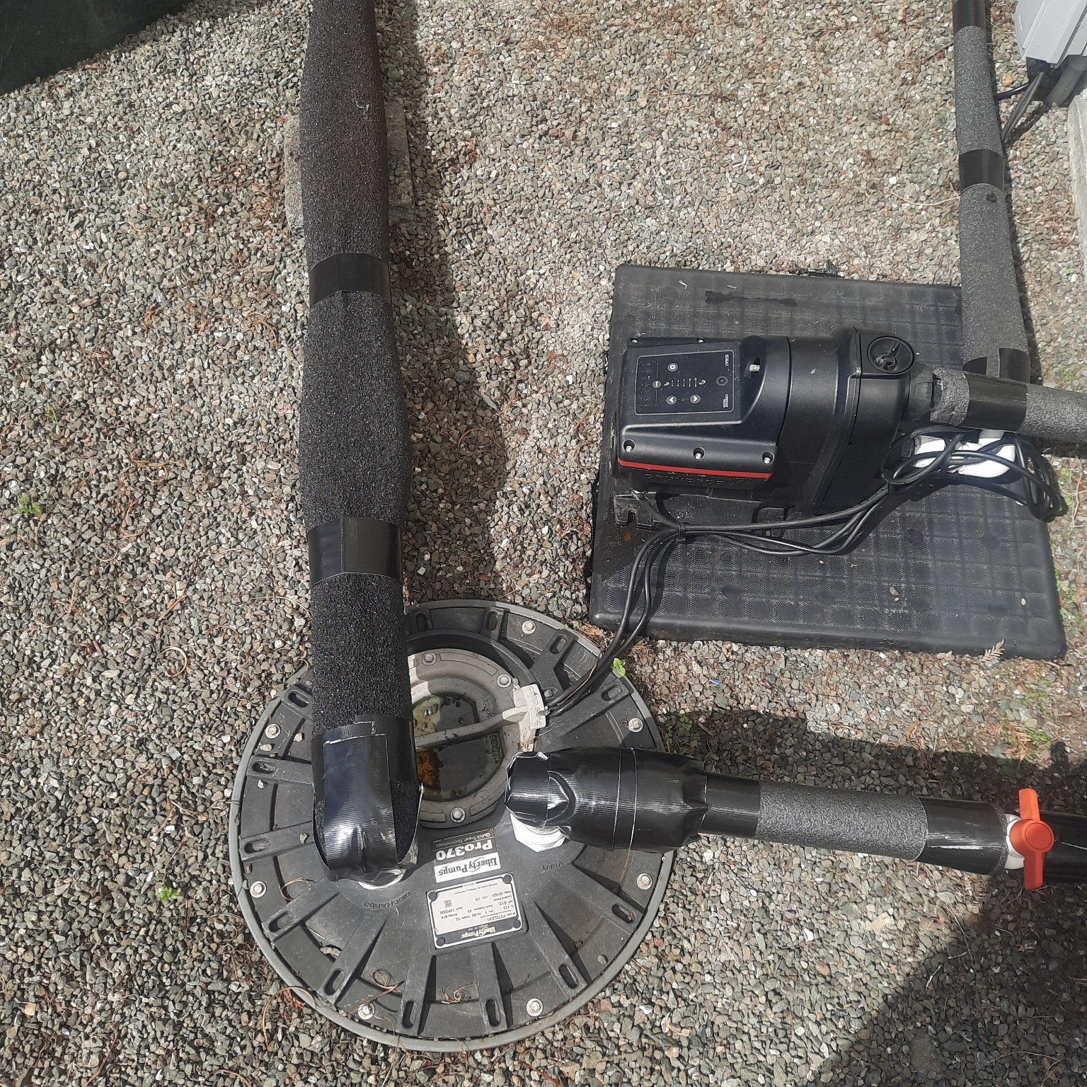 Above-ground booster pump and controller on a mat beside a round buried-sump access lid, with insulated pipe runs on gravel