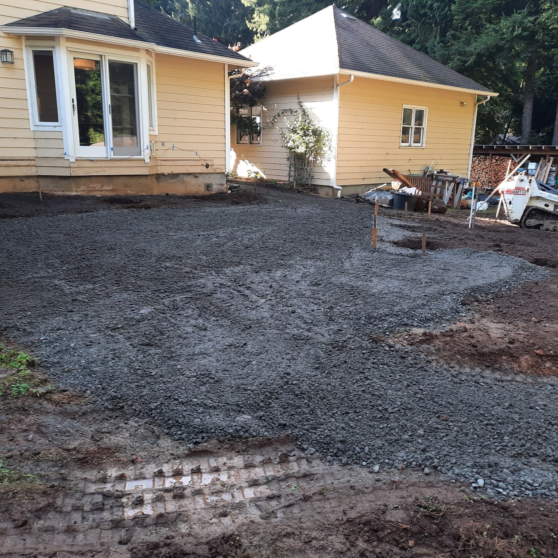Graded and compacted base material behind the house before deck construction, with layout stakes and a compact track loader on site