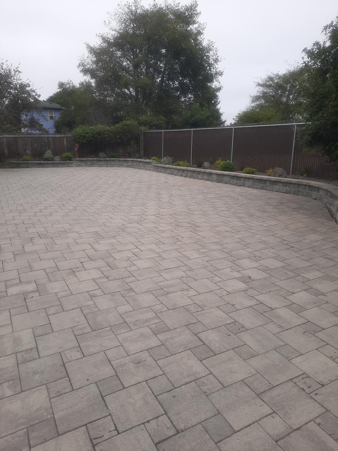Photograph of an open paver driveway bordered by a low block wall, fencing, and trees.
