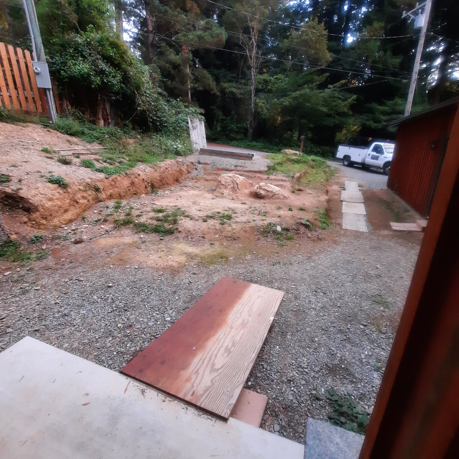 Photograph looking out from a doorway over bare graded soil, gravel, and a board walkway toward a wooded road.