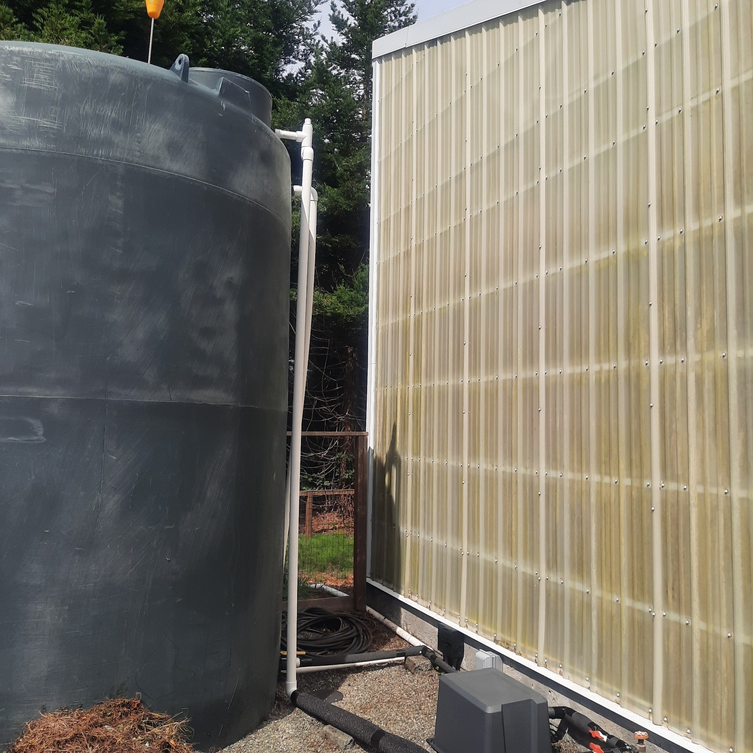 Large dark rainwater storage tank beside a translucent greenhouse wall, with white and black piping running along its base
