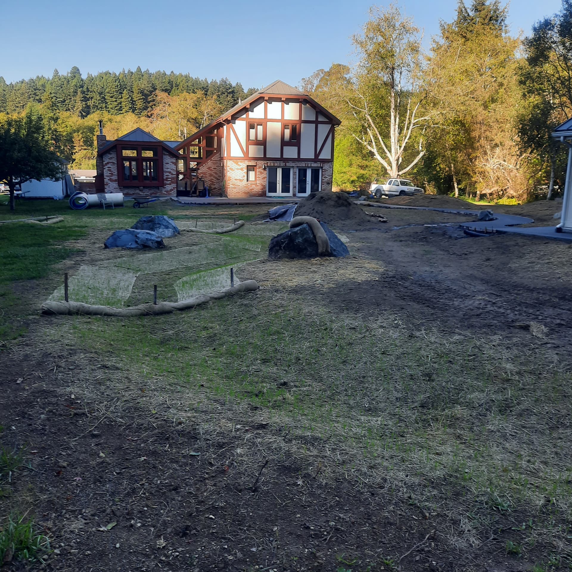 Property mid-construction with bare graded soil, erosion-control wattles, staged boulders, and a new curved path behind a large house