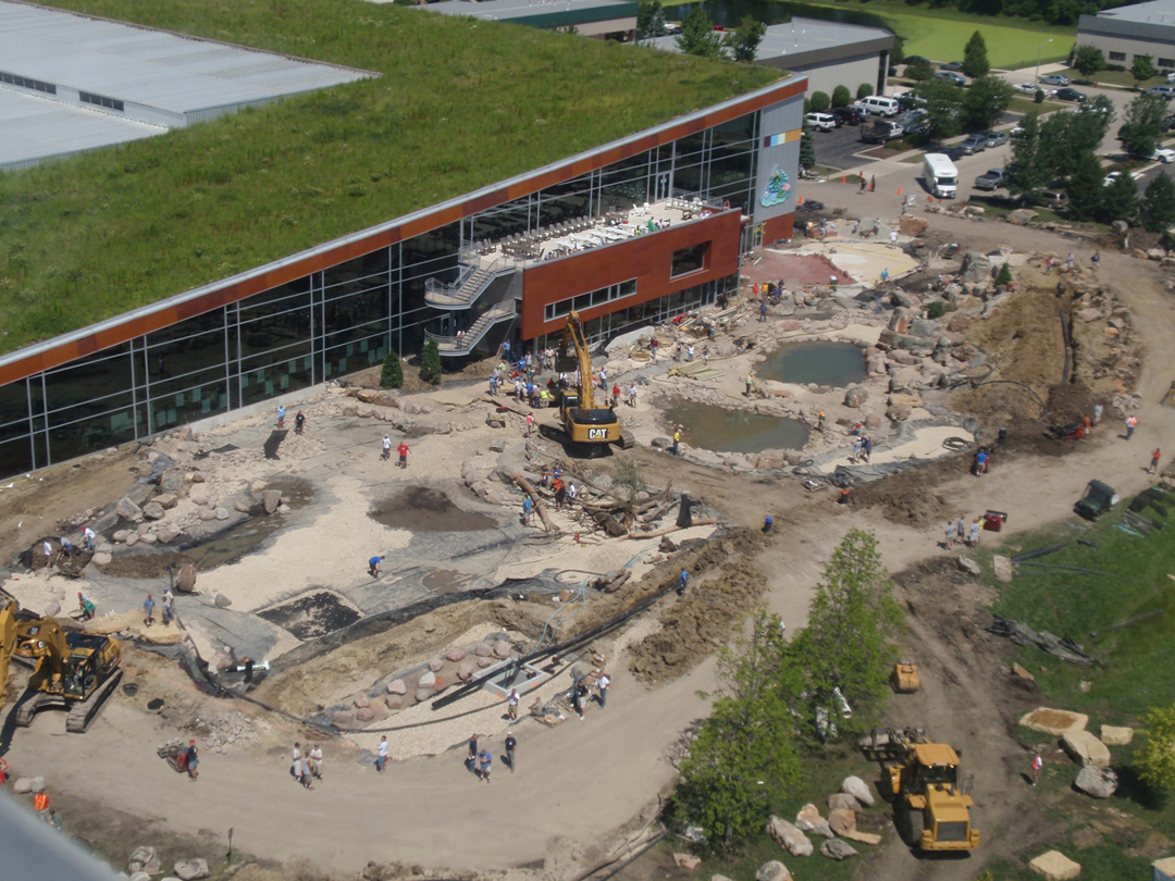 Overhead mid-construction view of Aquascape's large Aqualand water feature showing excavation, liner, stone placement, equipment and crews working across the site.