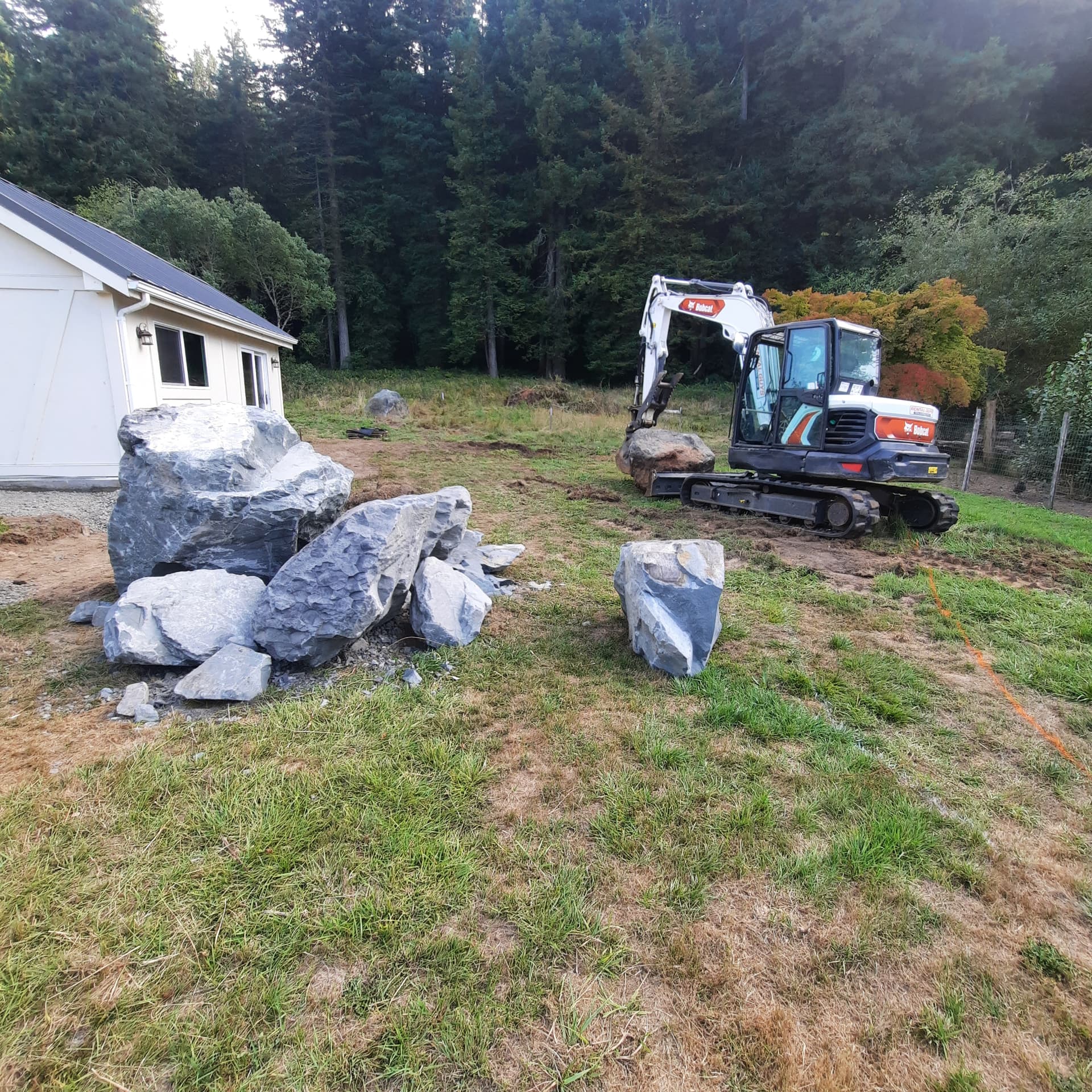 Compact excavator setting large stone boulders on a graded site beside a white outbuilding