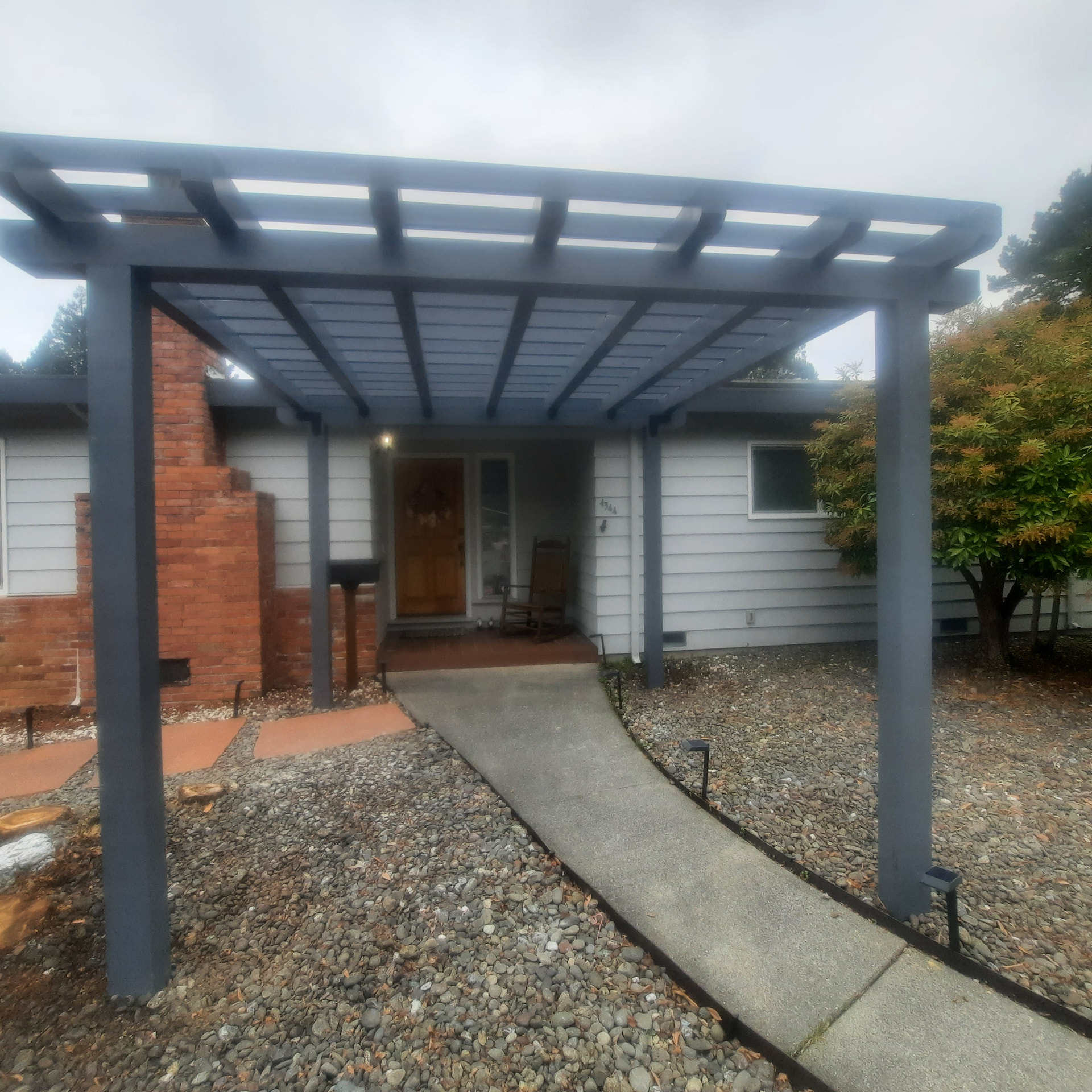 Gray entry pergola extending over a curved path beside a red brick chimney.