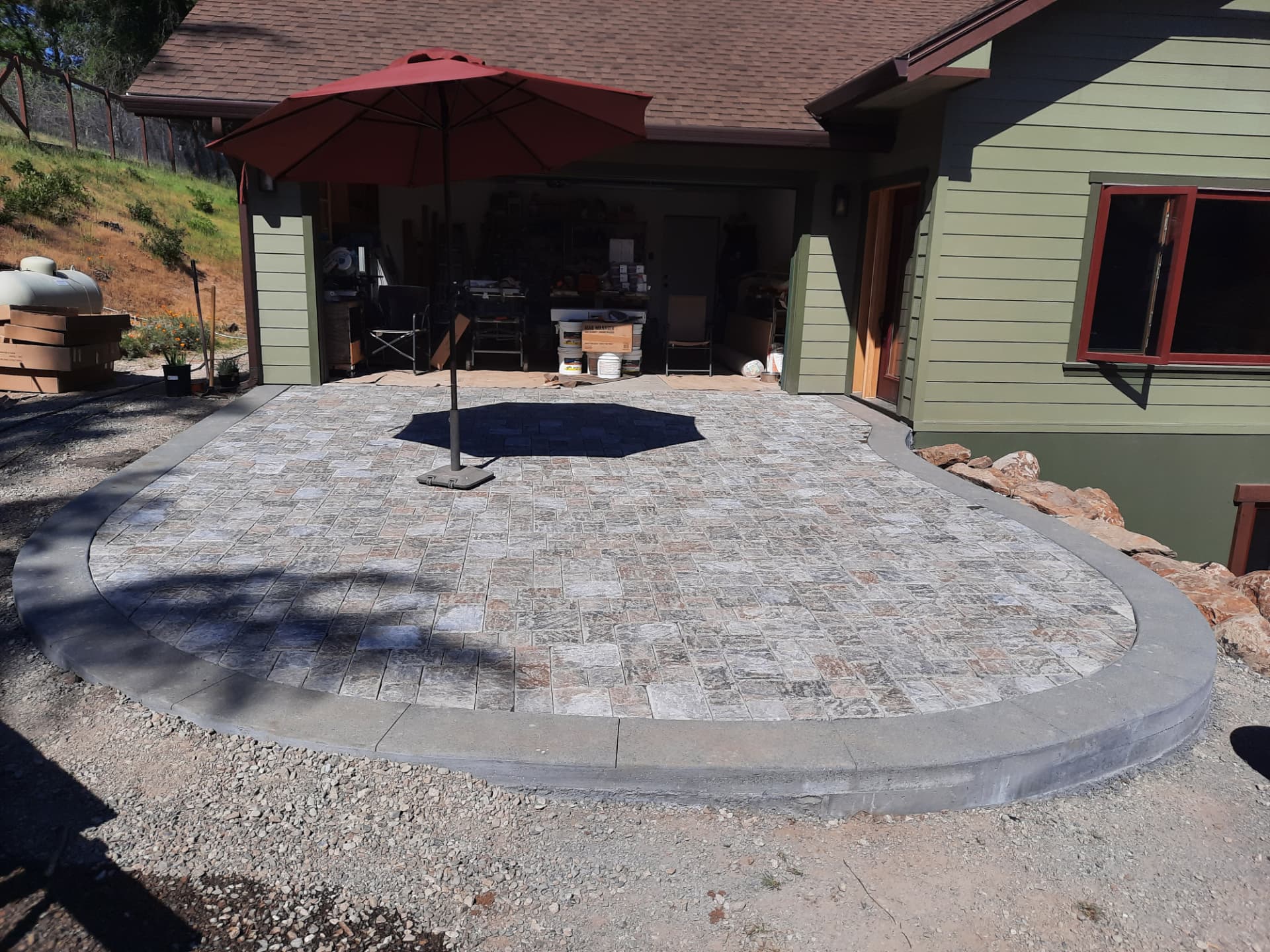 Curved paver patio with a stone border and umbrella at the front of a green house