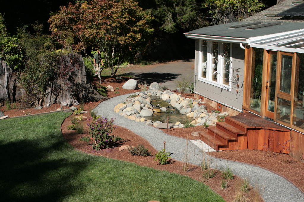 Living Earth landscape project with a small pond beside a home, curved gravel path, planting beds, lawn and wood deck.