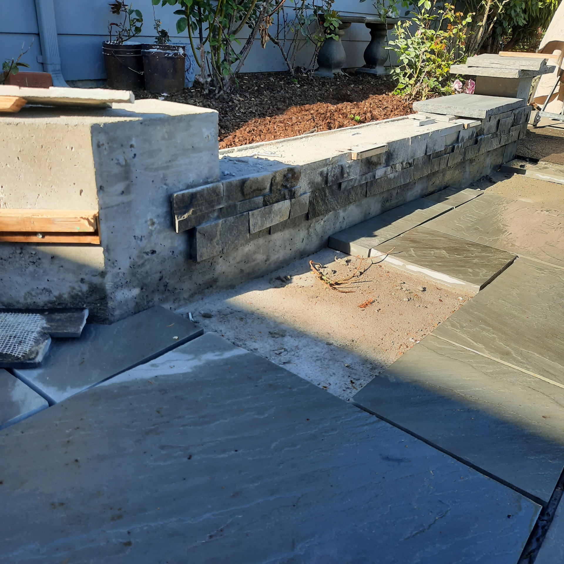 Retaining and planter walls under construction with stacked stone veneer over concrete and paving stones staged nearby